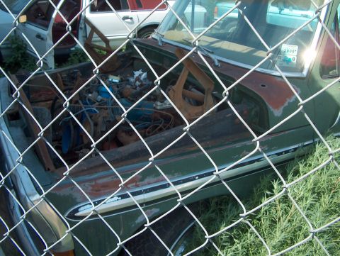 1979 Ford Bronco - A green 1978 Ford Bronco sits in a junkyard, its hood open to reveal the engine, partially obscured by a fence.