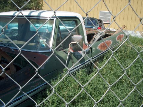 1979 Ford Bronco - A green pickup truck is seen parked behind a chain-link fence, with another vehicle visible in the background.