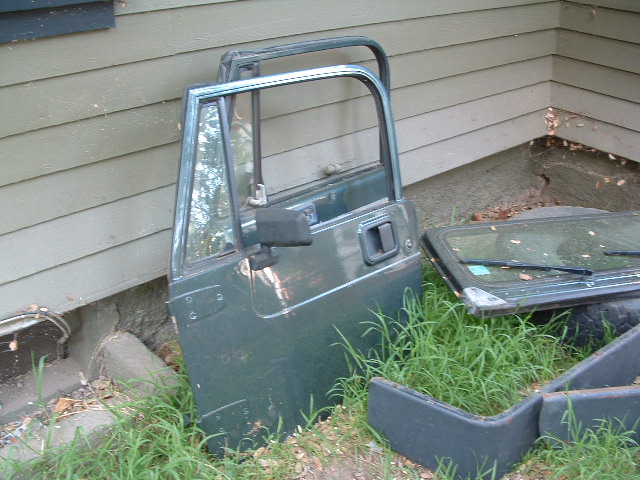 Green Jeep YJ door leaning against a wall, with a windshield frame and other parts on the ground