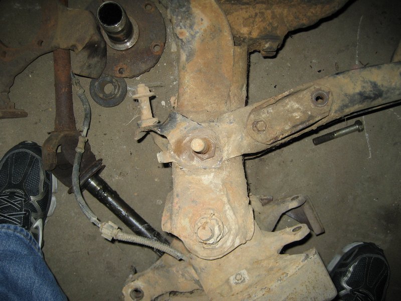 close-up of rusty suspension parts and a driveshaft with a person's foot in the corner