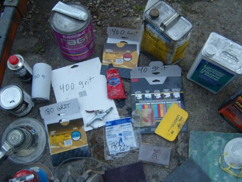 1989 Ford Bronco - Various automotive body repair supplies are laid out on the ground, including sandpaper, filler, and a sander.