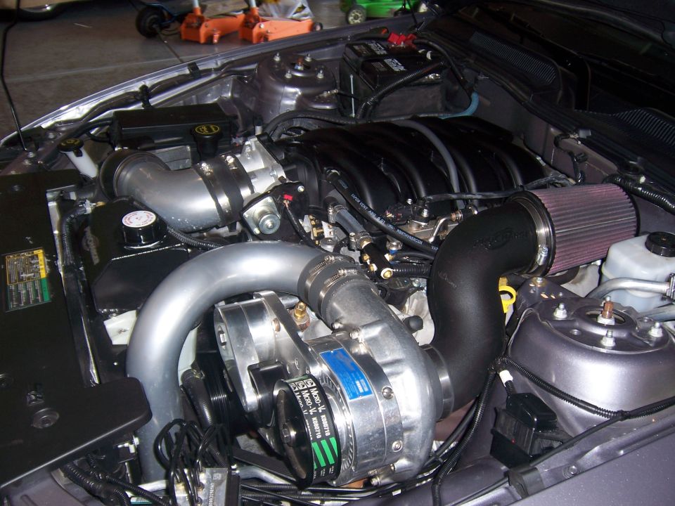 Engine bay with a polished supercharger, black intake piping, and a red air filter. The engine has black covers