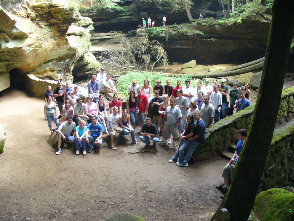 Large group of people gathered in a rocky, wooded area with a cave-like overhang