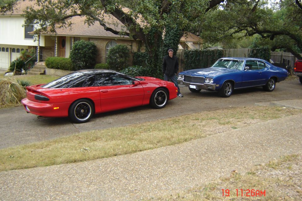 Red Camaro coupe and blue classic Buick two-door parked in a driveway with a man standing between them