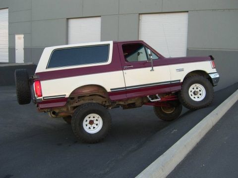 1991 Ford Bronco - A two-tone 1991 Ford Bronco is parked on an incline in front of a building with roll-up doors.