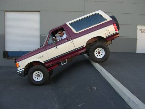 1991 Ford Bronco - A two-tone 1991 Ford Bronco is shown with its front wheels on a concrete barrier, demonstrating its lifted suspension.