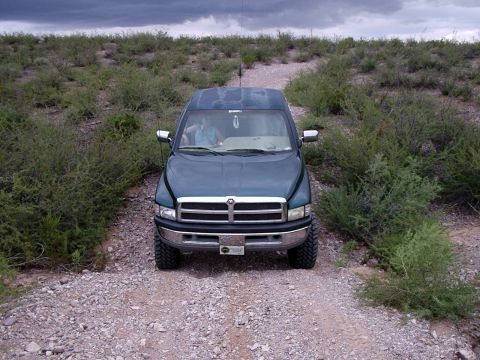 1996 Ford Bronco - A pickup truck drives down a rocky path surrounded by arid vegetation under a cloudy sky.