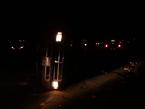 1996 Ford Bronco - A vehicle lies on its side at night, its hazard lights illuminated against the dark.