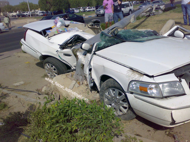 White sedan, heavily damaged, front end crushed, driver's side door open, windshield cracked, resting on curb