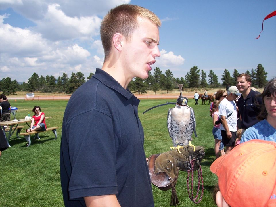 Guy in navy polo shirt holding a falcon on his gloved hand. People and trees in background