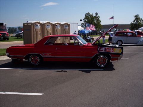 1972 Ford Ranchero - A red muscle car with a prominent 442 decal is displayed at an outdoor event, with other vehicles and portable toilets visible.