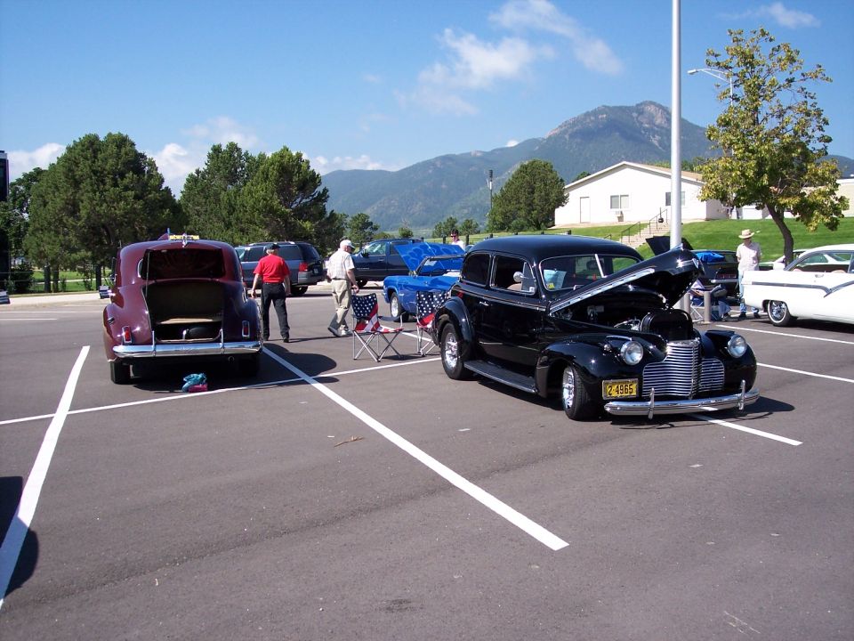 Black sedan with hood up, parked next to a maroon sedan with trunk open. Mountains in background