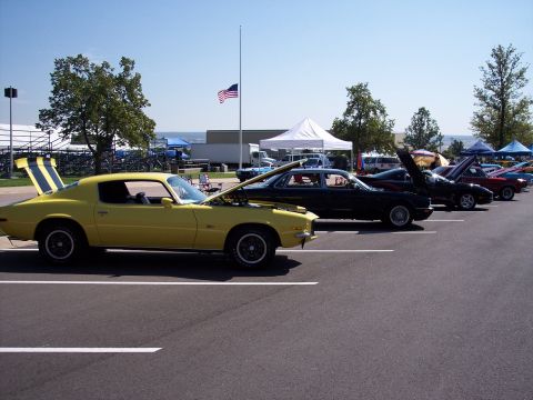 1972 Ford Ranchero - A line of cars are parked in a lot under a clear blue sky, with an American flag flying at half-mast.