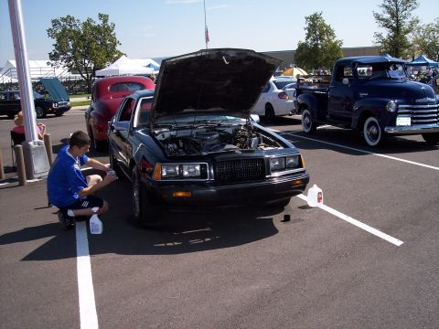 1972 Ford Ranchero - A person cleans the wheel of a black car with its hood open, parked next to other vehicles.