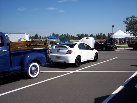 1972 Ford Ranchero - A white sedan with a spoiler and black rims is parked at an outdoor event with its hood open.