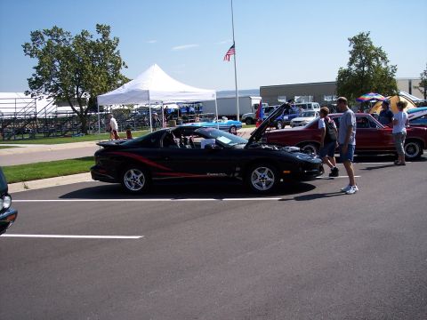 1972 Ford Ranchero - A black Pontiac Firebird with red racing stripes is parked at an outdoor event, with other vehicles and tents in the background.