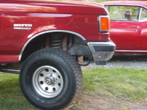 1989 Ford Bronco - Close-up of a red Ford Bronco's front wheel and fender, with another red vehicle visible in the background.