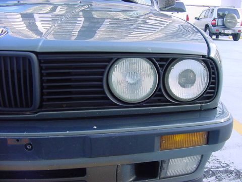 1989 BMW 3 Series - Close-up of the front of a 1989 BMW 3 Series, showing the grille and headlights. An SUV is visible in the background.