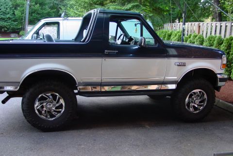 1993 Ford Bronco - A two-tone 1993 Ford Bronco with chrome wheels is parked on asphalt, with a white van visible behind it.