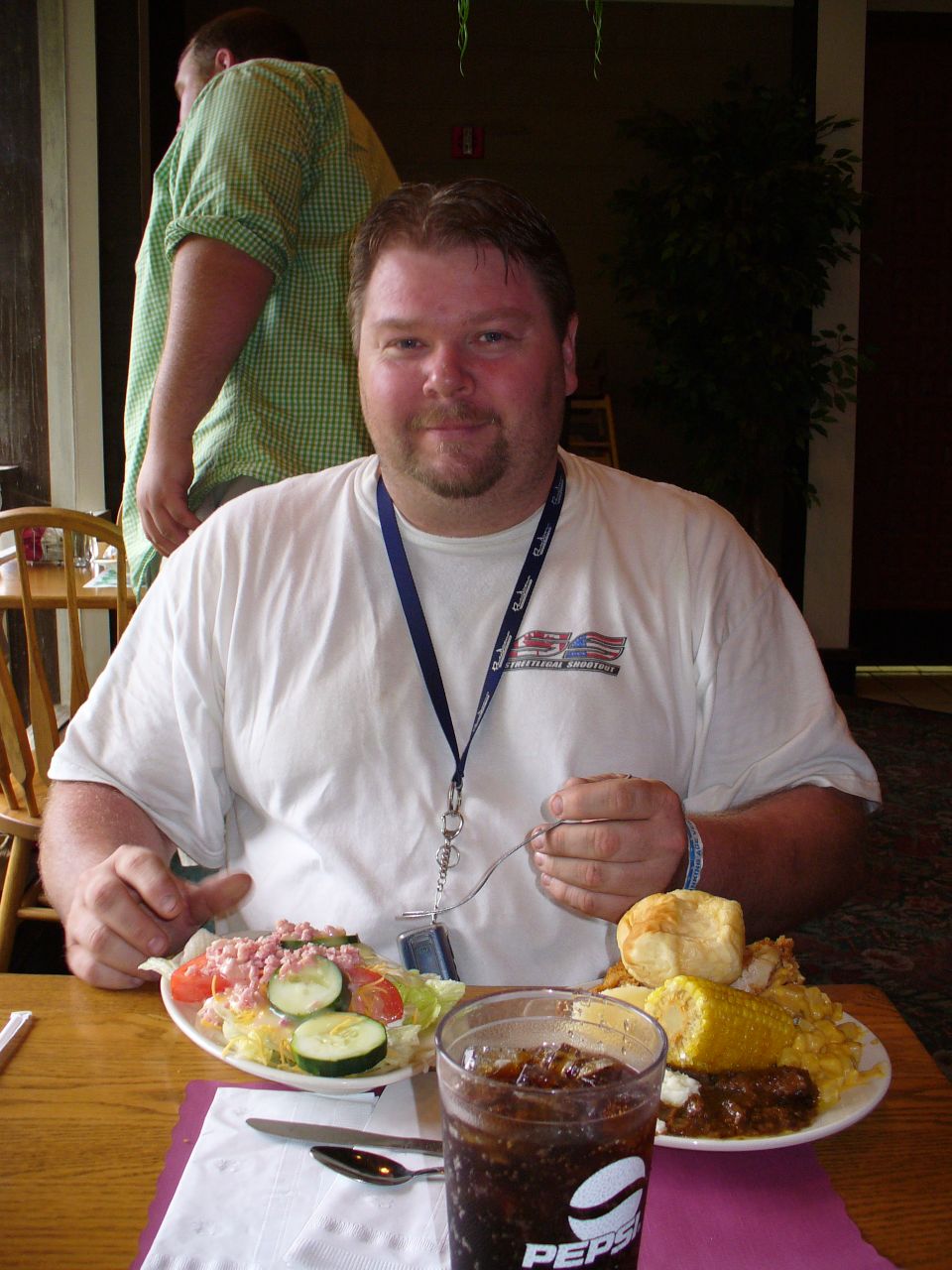Man in white t-shirt with lanyard eating a meal at a table with salad, corn, and a drink