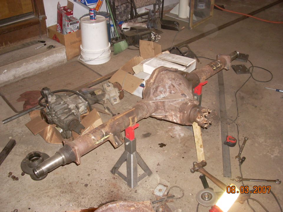 Vehicle rear axle assembly on jack stands in a garage. A transfer case is also visible
