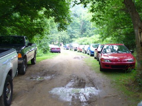 1991 Ford Probe - A line of cars is parked on a dirt road, with trees overhead and puddles on the ground.