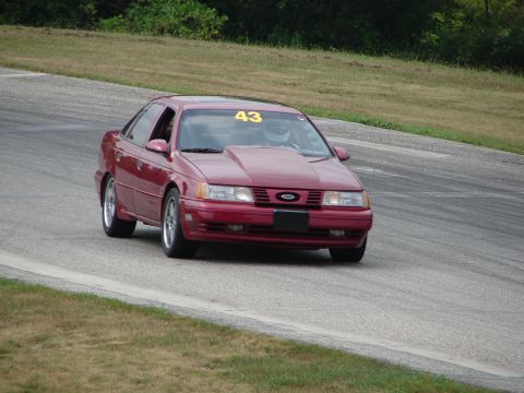 1993 Ford Taurus - A red 1993 Ford Taurus with a racing number on the windshield navigates a turn on a paved track.