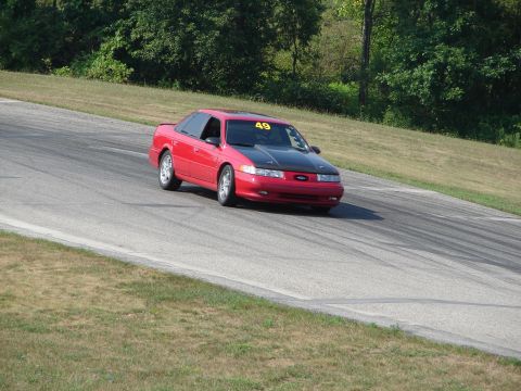 1993 Ford Taurus - A red 1993 Ford Taurus with a black hood navigates a turn on a road course, displaying the number 49 on its windshield.