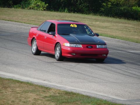 1993 Ford Taurus - A red 1993 Ford Taurus with a black hood is on a race track, displaying the number 49 on its windshield.