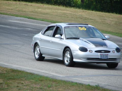 1993 Ford Taurus - Silver sedan with racing stripes and number 50 on windshield navigates a turn on a road course.