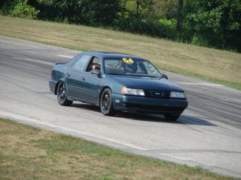 1993 Ford Taurus - A dark green sedan navigates a turn on a race track, displaying the number 54 on its windshield.