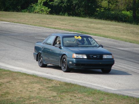 1993 Ford Taurus - A dark green sedan with a racing number on its windshield navigates a turn on a paved track bordered by grass.