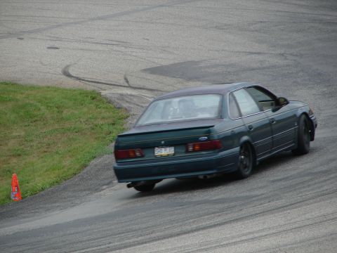 1993 Ford Taurus - A dark green sedan drifts around a corner on a race track, its rear end angled towards the camera.