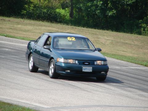 1993 Ford Taurus - A dark green sedan with racing number 62 on the windshield drives on a paved track with grassy shoulders.