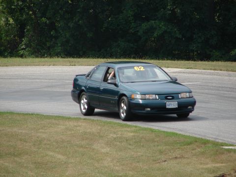 1993 Ford Taurus - A dark green sedan with racing number 62 on the windshield navigates a turn on a road course.