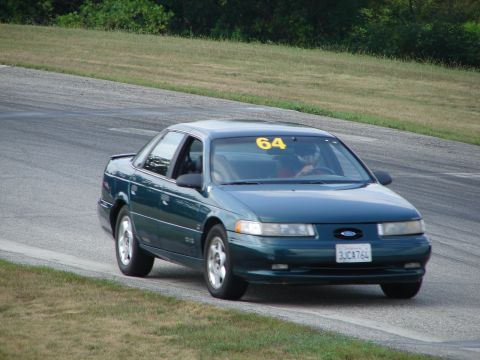 1993 Ford Taurus - A dark green sedan navigates a turn on a paved track, with the number 64 displayed on its windshield.