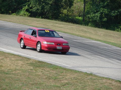 1993 Ford Taurus - A red sedan with racing number 69 navigates a turn on a paved road course with grassy verges and trees in the background.