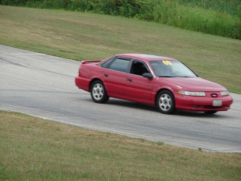 1993 Ford Taurus - Red 1993 Ford Taurus driving on a road course with a racing number on the windshield.