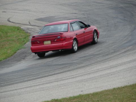 1993 Ford Taurus - A red 1993 Ford Taurus sedan navigates a turn on a race track, leaving tire marks on the asphalt.