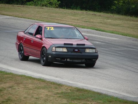 1993 Ford Taurus - A red sedan, identified as a 1992 model, navigates a turn on a road course with a racing bra covering its front end.