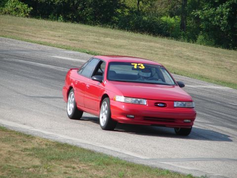1993 Ford Taurus - A red 1993 Ford Taurus navigates a turn on a paved race track with grassy shoulders.
