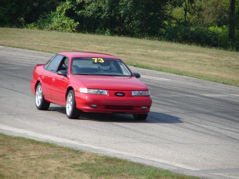 1993 Ford Taurus - A red sedan with racing number 73 on the windshield navigates a turn on a road course.