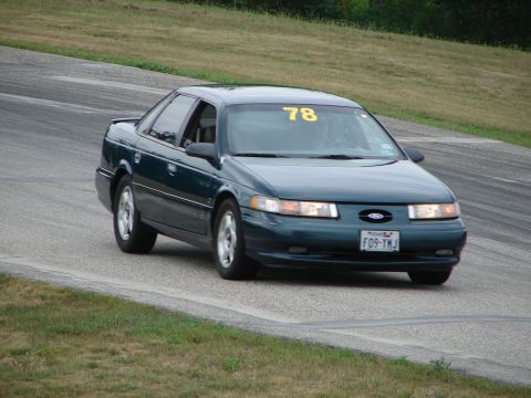 1993 Ford Taurus - Dark green 1993 Ford Taurus driving on a road course with racing numbers on the windshield.