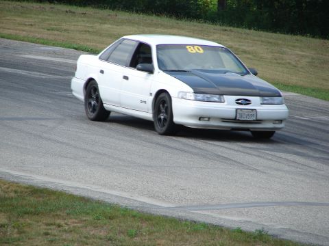 1993 Ford Taurus - A white sedan with a black hood navigates a turn on a race track, displaying the number 80 on its windshield.