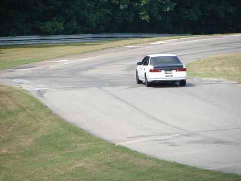 1993 Ford Taurus - A white 1993 Ford Taurus sedan drives on a paved road course with green grass and trees in the background.