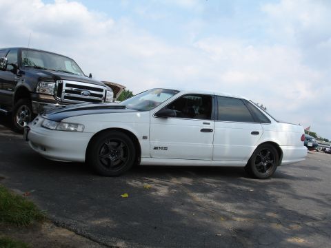 1993 Ford Taurus - A white sedan with a contrasting black hood is parked next to a large truck under a cloudy sky.