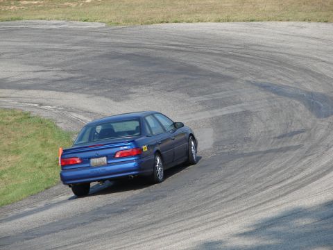 1993 Ford Taurus - A blue sedan navigates a turn on a paved race track, with grass and trees visible in the background.
