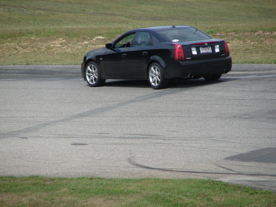 Black sedan on asphalt with grass in background. Rear view shows taillights and dual exhaust