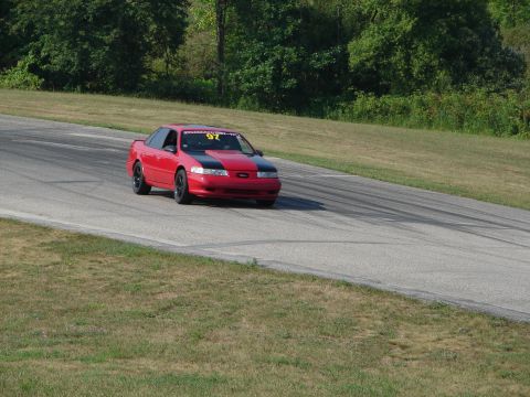 1993 Ford Taurus - A red coupe with a black hood and racing number 97 drives on a paved track surrounded by grass and trees.