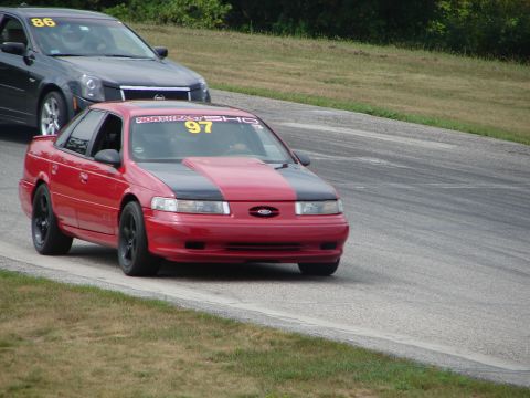 1993 Ford Taurus - A red 1993 Ford Taurus SHO with a black hood stripe and racing numbers competes on a road course, followed by another vehicle.